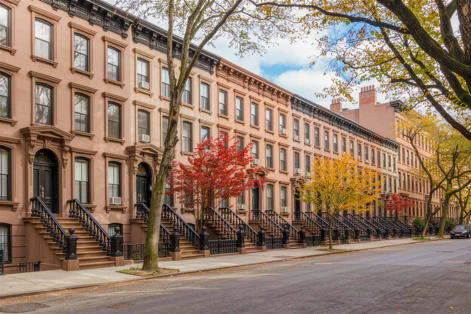 A block of brownstone row houses with stoops and street trees under a blue autumn sky