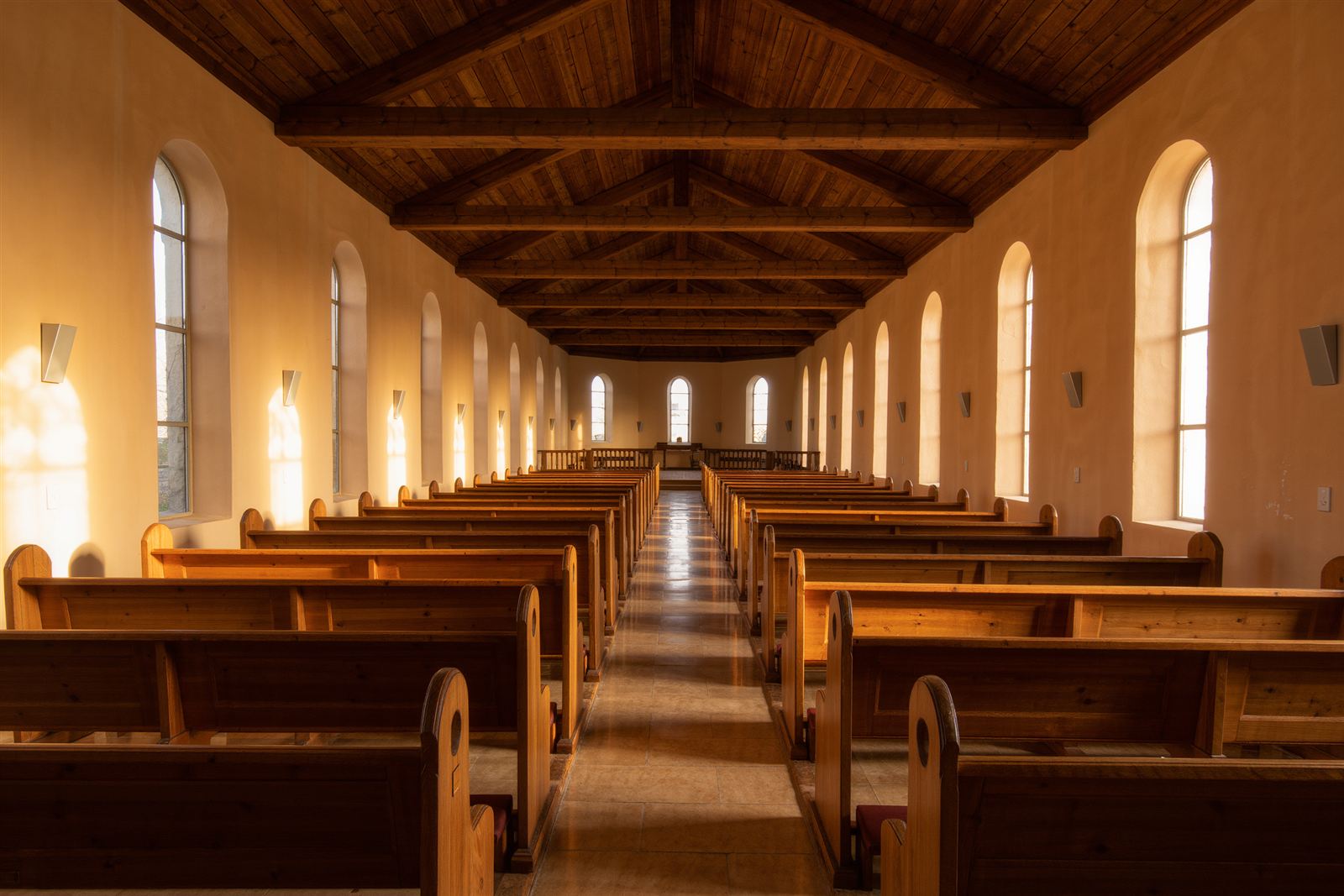 Rows of empty wooden pews in a sunlit church with tall arched windows